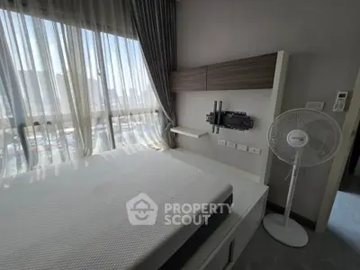Modern bedroom with large window and city view, featuring a wall-mounted TV bracket and standing fan.