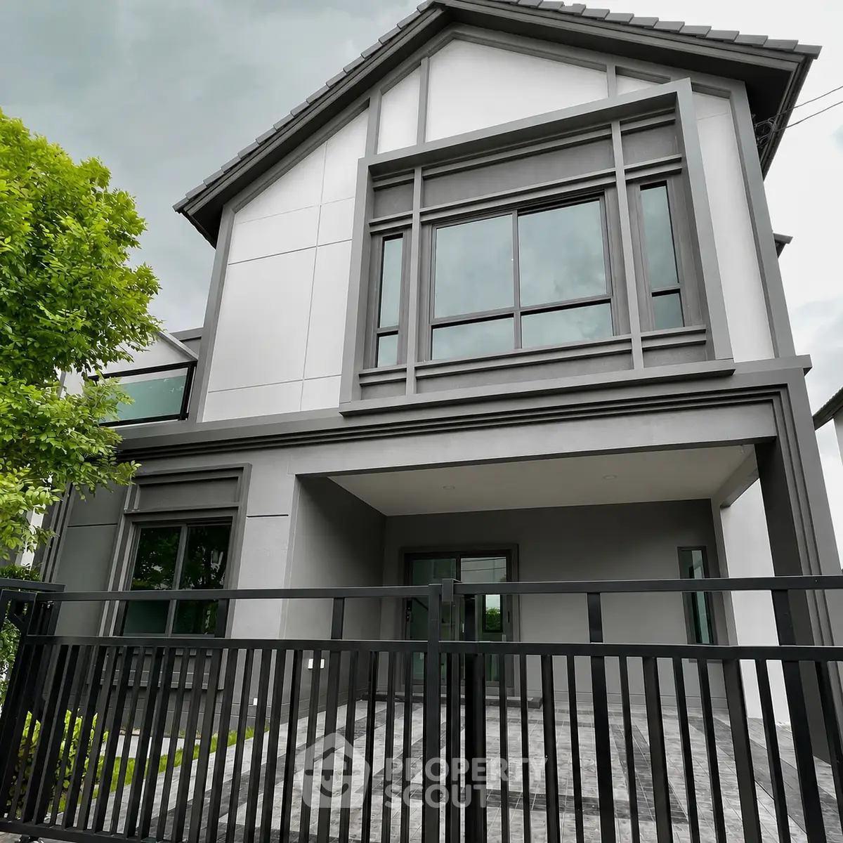 Modern two-story house with sleek design and large windows, featuring a gated entrance and lush greenery.