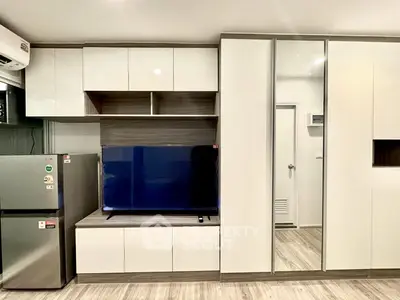 Modern apartment interior with sleek storage cabinets and a flat-screen TV, featuring a compact fridge and stylish flooring.