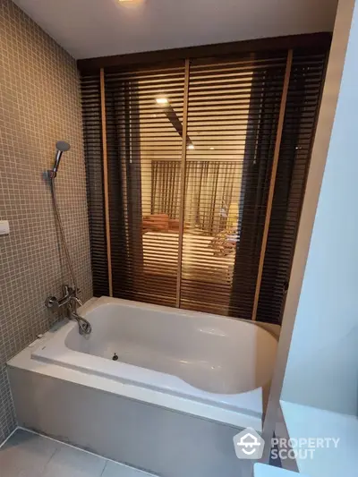 Elegant bathroom with a large bathtub, modern fixtures, and mosaic tile walls, complemented by warm wooden blinds and ambient lighting.