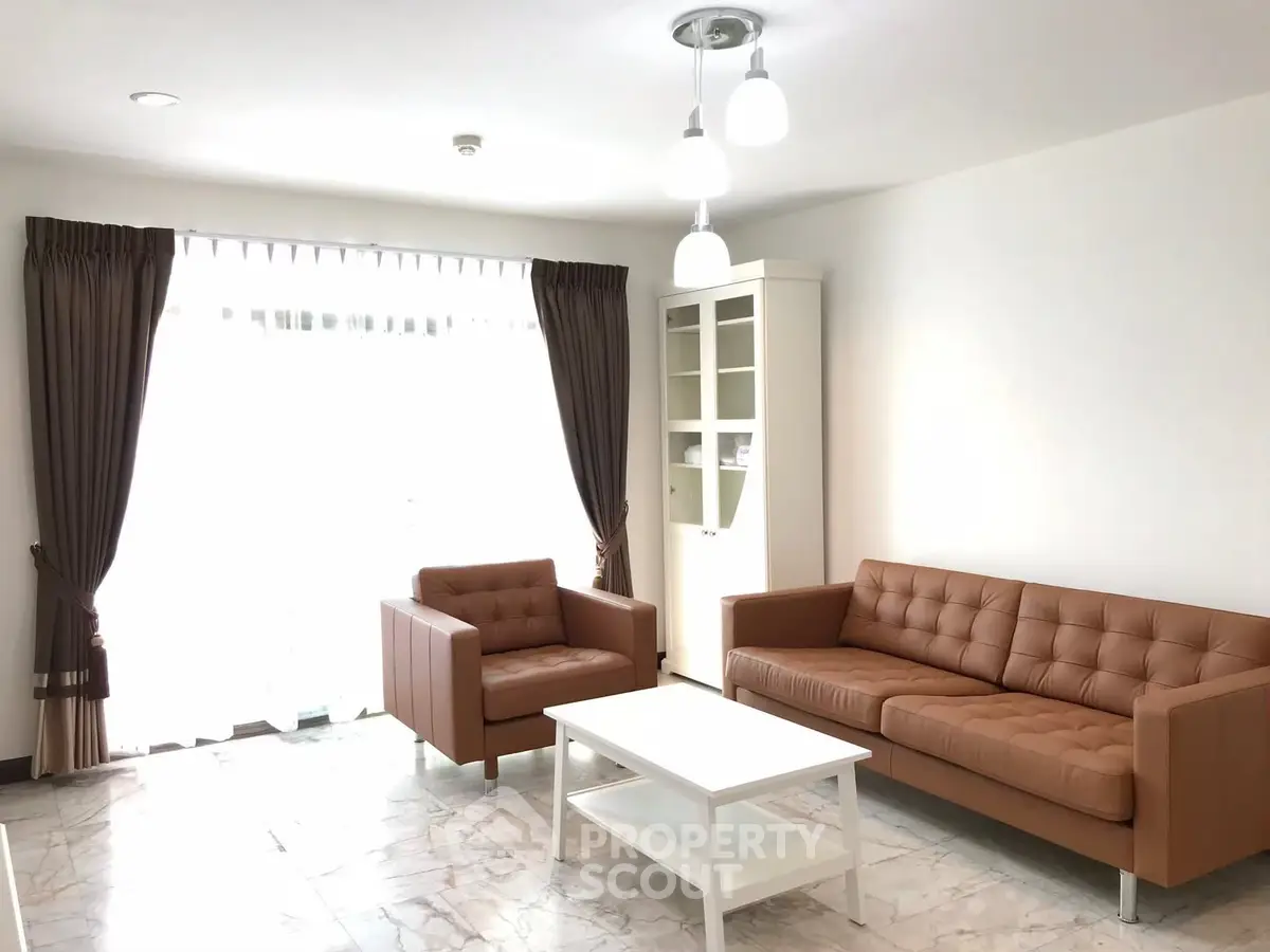 Bright living room with modern brown leather sofa and armchair, white coffee table, and large window.