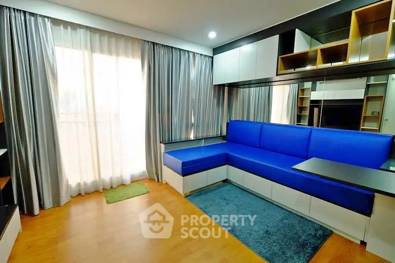 Modern living room with blue sofa, wooden flooring, and ample natural light from large window.