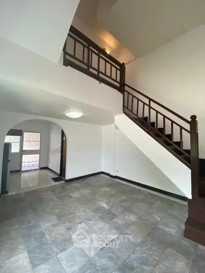 Spacious living area with elegant marble flooring and a grand wooden staircase leading to an upper level, featuring ample natural light and a unique arched doorway.