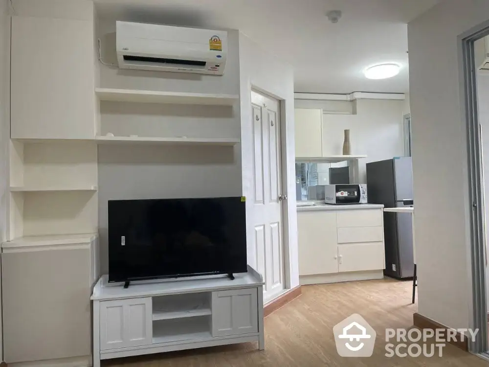 Modern apartment interior with TV and kitchen area, featuring sleek white cabinetry and wood flooring.