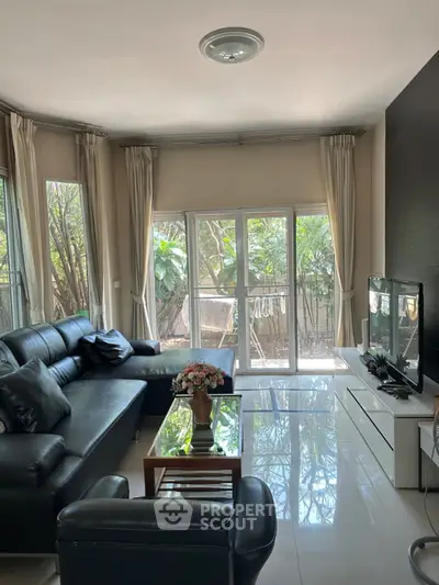 Spacious living room with modern black leather sofa and large windows overlooking lush greenery.