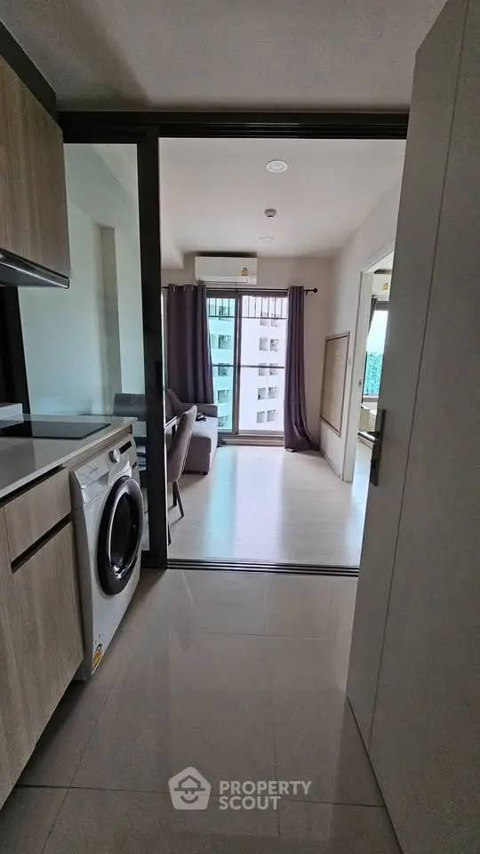 Modern apartment interior with kitchen and living room, featuring washing machine and large windows.