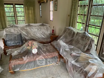 Spacious living room with vintage furniture covered in protective plastic, large windows, and garden view.