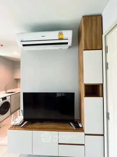Modern living room with sleek TV unit and air conditioning, featuring a compact kitchen with washing machine.