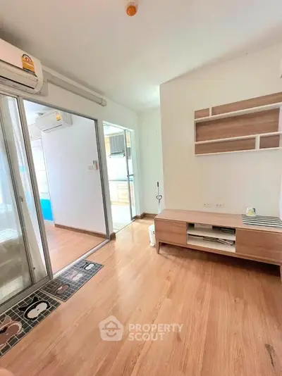 Modern living room with wooden flooring and built-in shelves, featuring a sliding glass door leading to a bright kitchen.