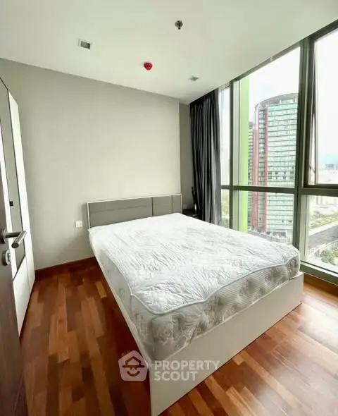 Modern bedroom with large windows and city view, featuring a comfortable bed and wooden flooring.