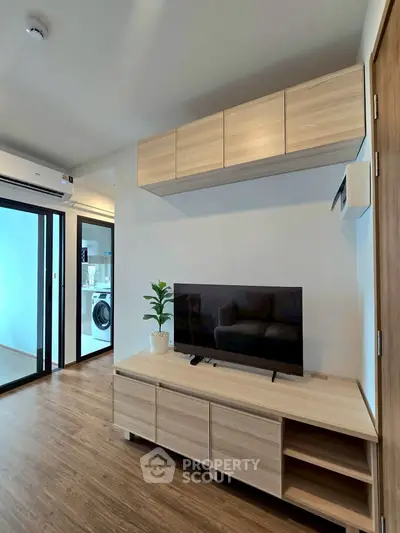 Modern living room with sleek wooden furniture and a flat-screen TV, adjacent to a laundry area with a washing machine.