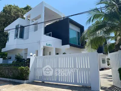 Modern white villa with sleek design and lush greenery, featuring a gated entrance and large windows.
