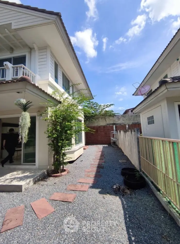 Charming two-story house with a spacious gravel pathway and lush greenery under a bright blue sky.