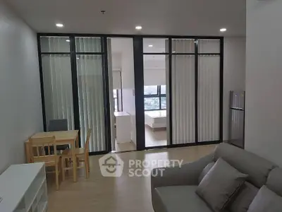 Modern open-plan living room with dining area and view into bedroom, featuring sleek furniture and large windows.