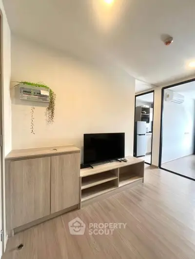 Modern living room with wooden flooring and sleek TV stand, leading to a kitchen area with appliances.