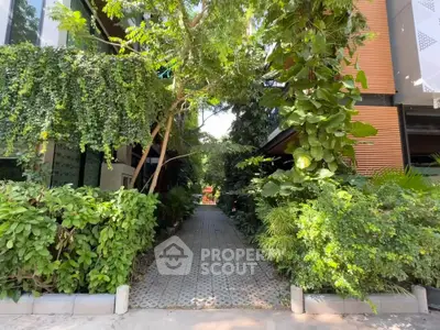 Lush green pathway leading to a modern building entrance, surrounded by vibrant foliage.