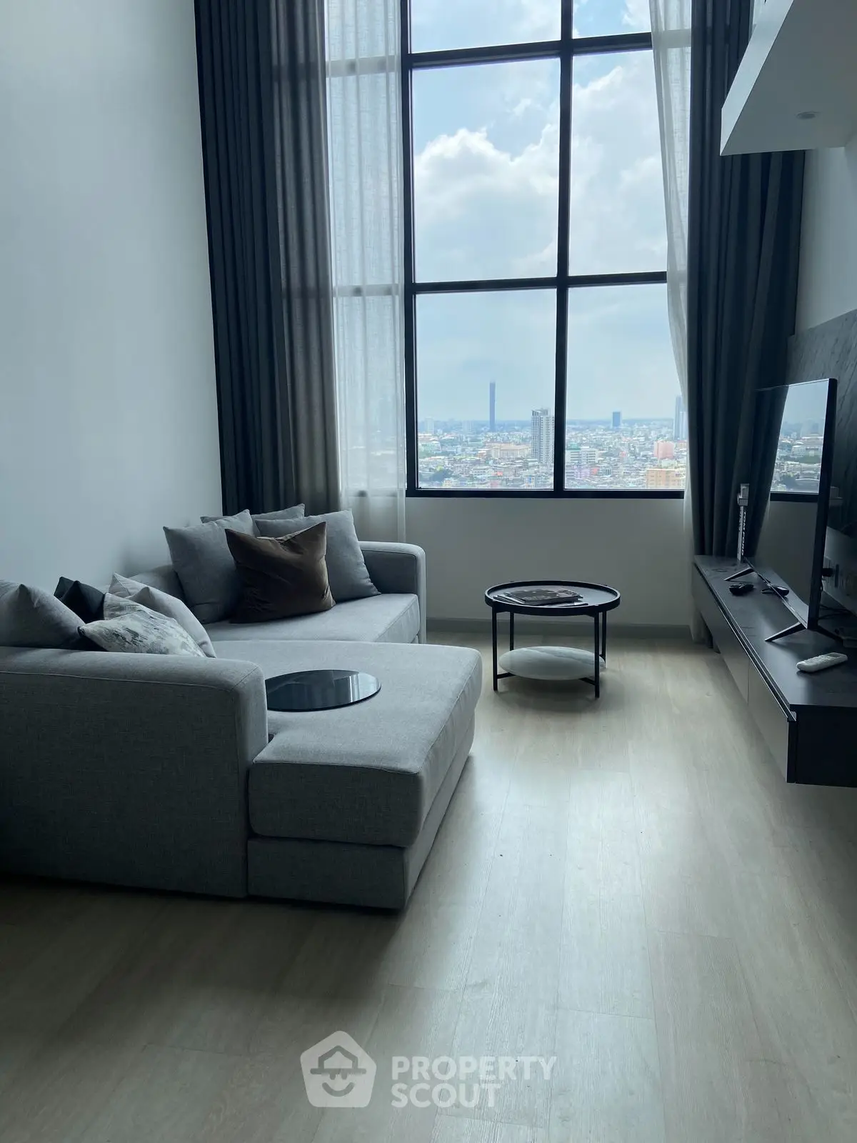 Modern living room with large window and city view, featuring a stylish gray sofa and sleek TV setup.