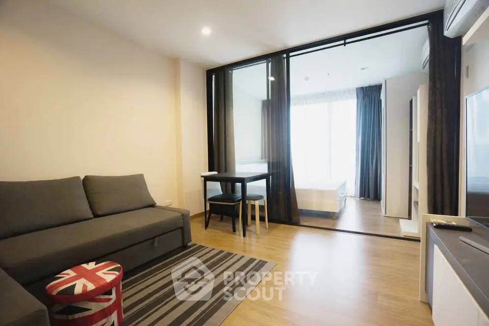 Modern living room with glass partition leading to bedroom, featuring stylish furniture and wooden flooring.
