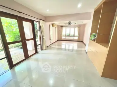 Spacious living room with large windows and garden view, featuring built-in shelves and elegant tile flooring.