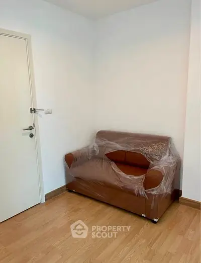 Cozy small living room with a brown sofa wrapped in plastic, wooden flooring, and a white door.