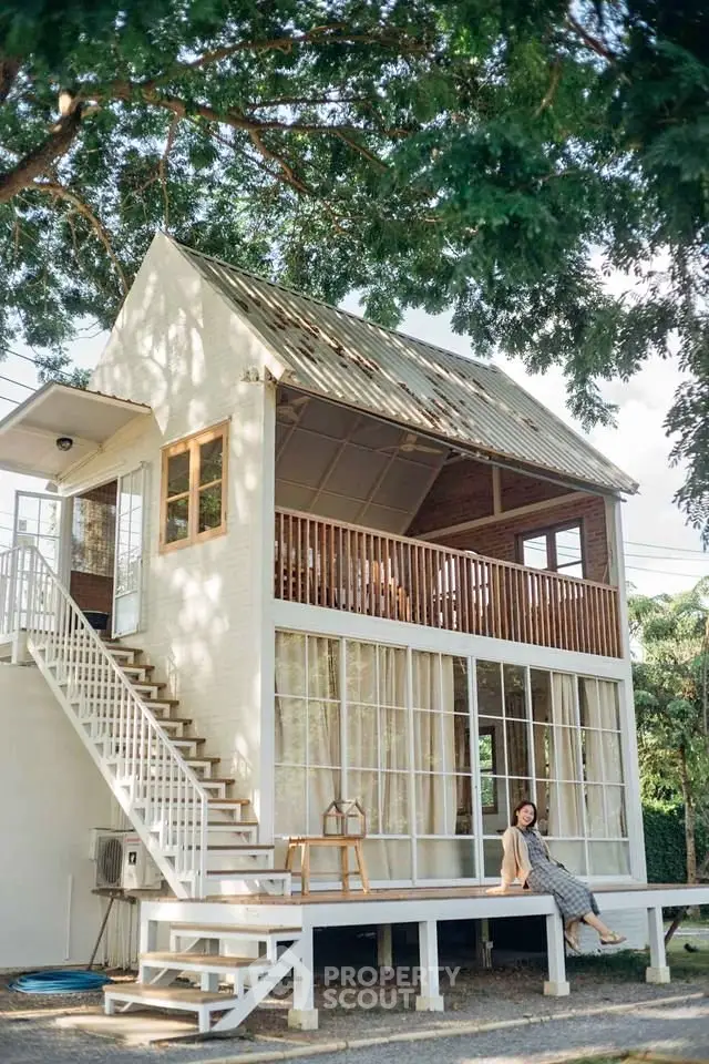 Charming two-story house with large windows and a spacious balcony under lush trees.