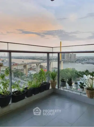 Stunning balcony view with lush plants overlooking a scenic river and bridge at sunset.