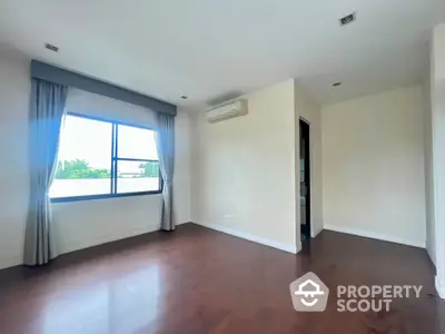 Spacious and well-lit living room with polished wooden flooring, large windows with blue curtains, and a clean white wall finish.