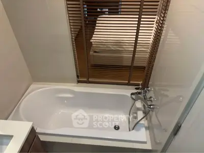 Modern bathroom with bathtub and view into cozy bedroom through blinds.