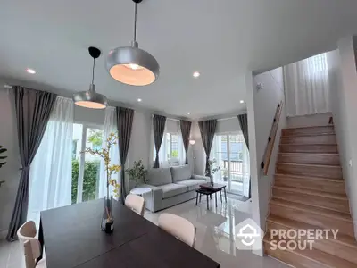 Modern living room with elegant dining area and staircase, featuring stylish lighting and natural light.