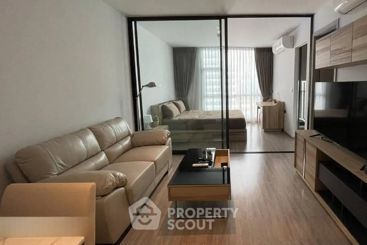 Modern living room with glass partition leading to a stylish bedroom, featuring sleek furniture and natural light.