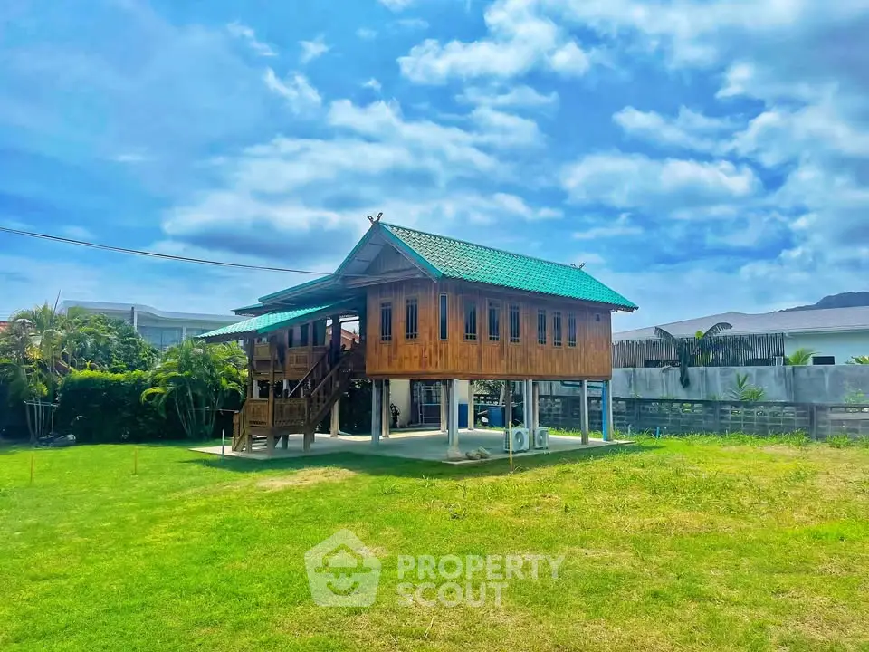 Charming elevated wooden house with green roof in lush garden setting under blue skies.