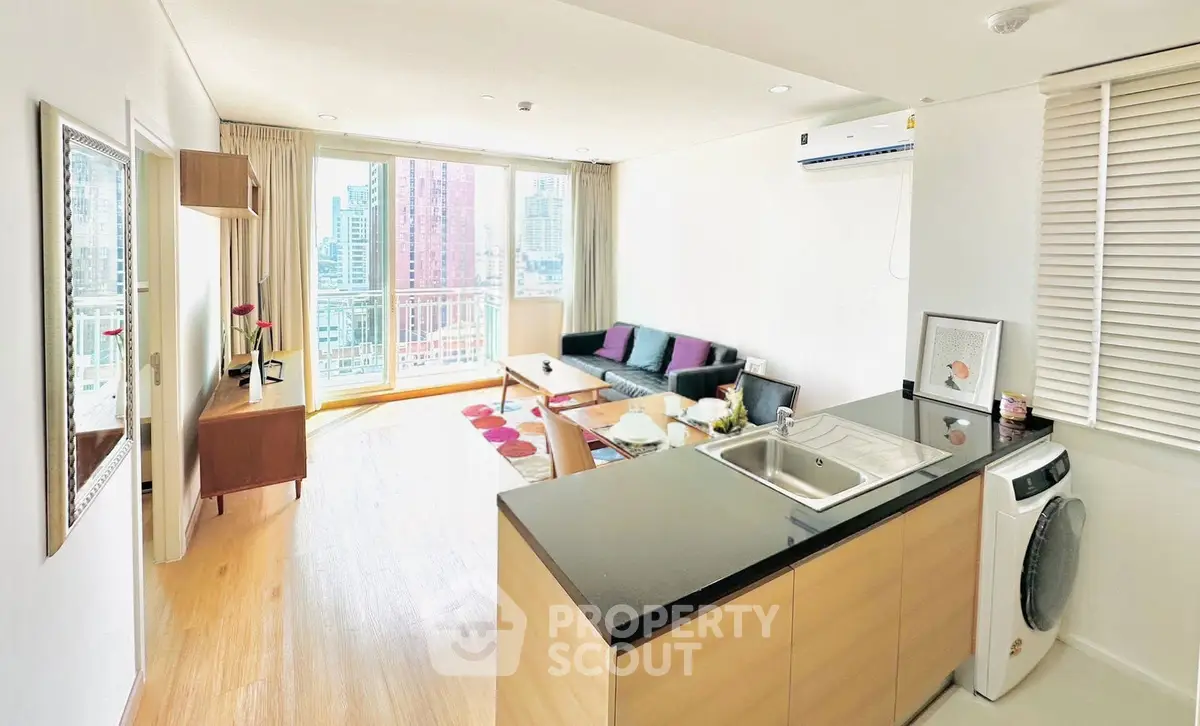 Bright modern living room with open kitchen and city view, featuring sleek furniture and natural light.