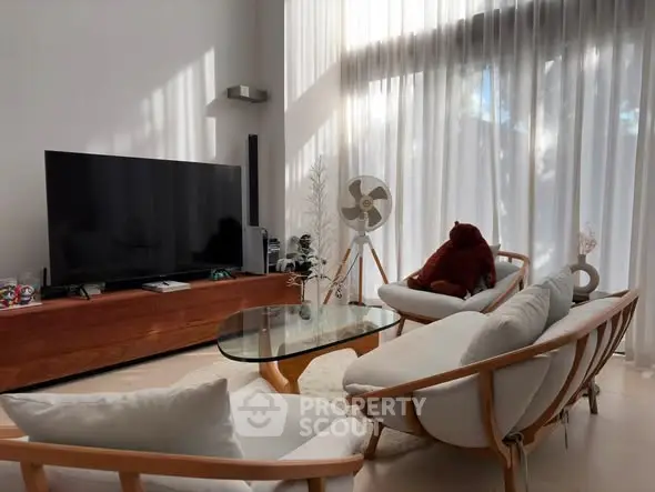 Stylish modern living room with large TV, elegant furniture, and natural light through sheer curtains.