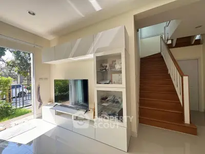 Bright living room with modern TV unit and staircase, featuring large windows and garden view.