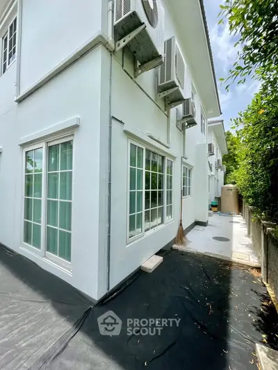 Modern white building exterior with large windows and air conditioning units, surrounded by greenery.