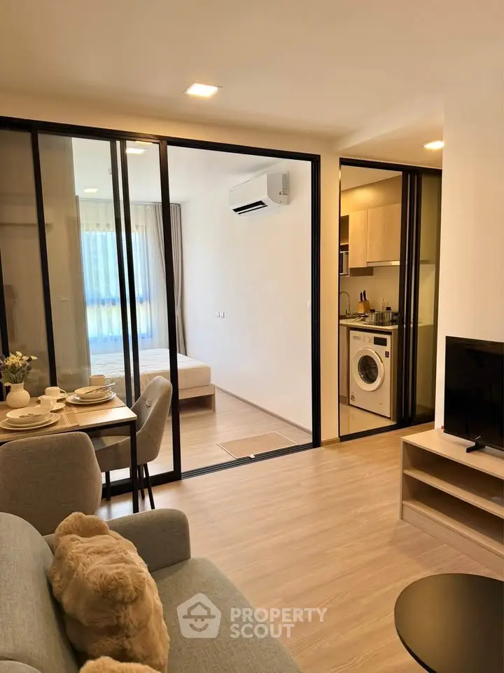 Modern open-plan living room with glass partition, cozy dining area, and compact kitchen featuring a washing machine.
