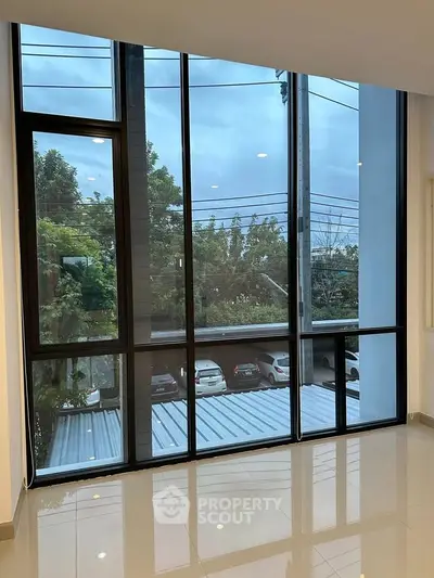 Spacious room with large floor-to-ceiling windows offering a serene view of trees and parked cars.