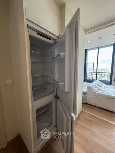 Modern apartment with open fridge and bedroom view, showcasing sleek design and natural light.