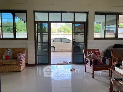 Spacious living room with large windows and view of driveway, featuring comfortable seating and tiled flooring.