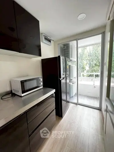 Modern kitchen with sleek cabinets, microwave, and fridge, leading to a bright balcony with greenery view.