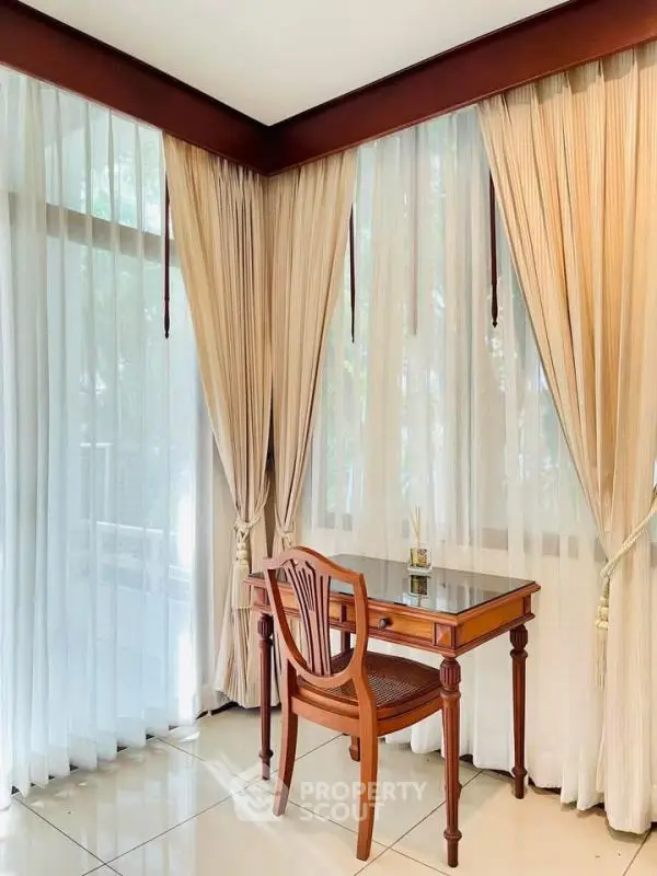 Elegant study nook with wooden desk and chair by large windows with sheer curtains.