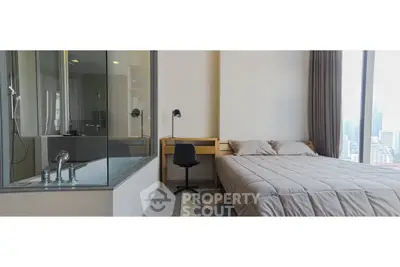 Modern bedroom with glass-enclosed bathroom and city view, featuring a cozy bed and study area.