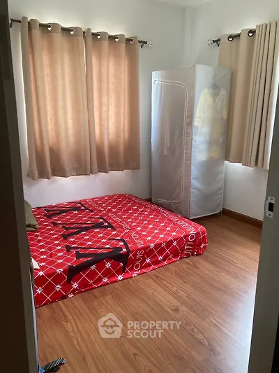 Cozy bedroom with red bedding and wooden flooring, featuring natural light from curtained windows.