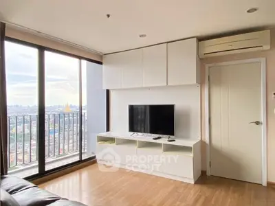 Modern living room with large windows and city view, featuring sleek white cabinetry and TV setup.