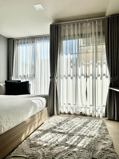 Bright and airy bedroom with large windows and elegant curtains, featuring a cozy bed and soft rug.
