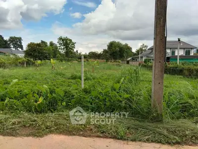 Spacious green land plot with potential for development, surrounded by lush vegetation and a nearby residential house.
