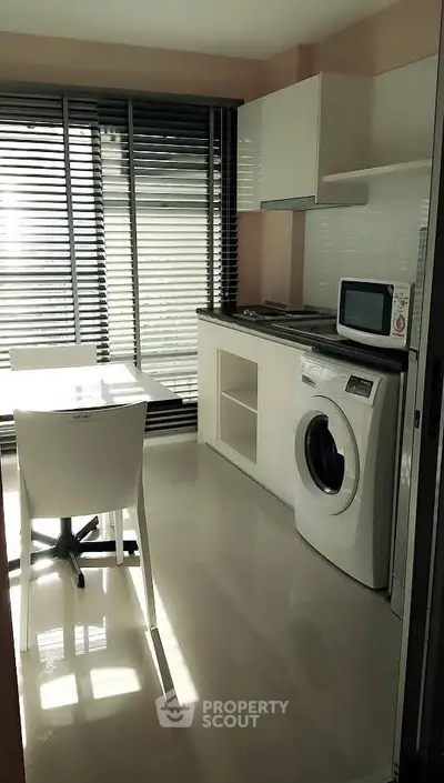 Modern kitchen with washing machine and microwave, featuring sleek blinds and a dining table.