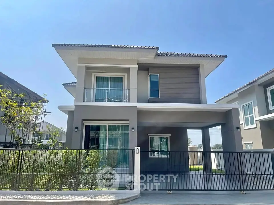 Modern two-story house with balcony and gated entrance under clear blue sky.