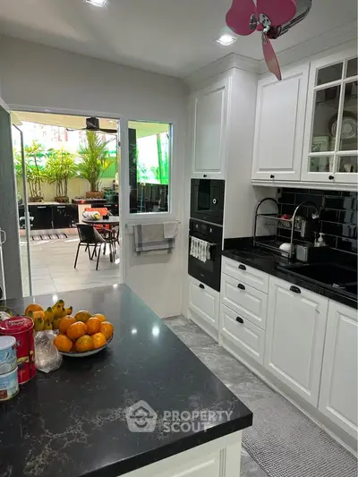Charming kitchen with black countertops and white cabinetry, opening to a lush outdoor dining area.