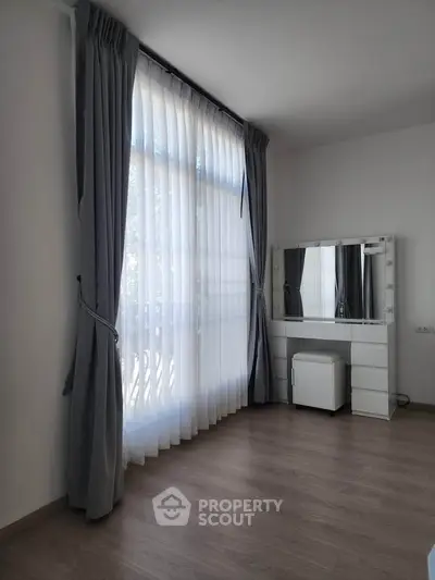 Bright bedroom with large window and elegant curtains, featuring a stylish vanity mirror and stool.